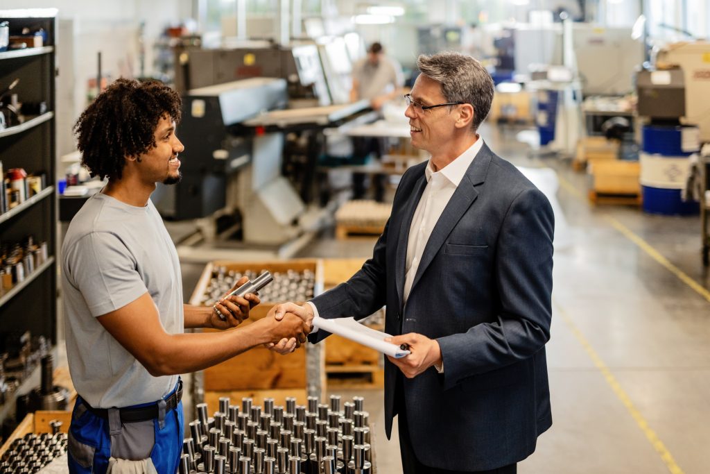 Geschäftsmann im Anzug schüttelt einem Werkstattmitarbeiter die Hand beim Verkauf von gebrauchtem Inventar